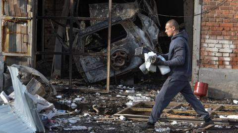 A man walks past a car repair workshop which was damaged by a Russian drone strike. The floor is covered in debris and there's a burnt out car in the background