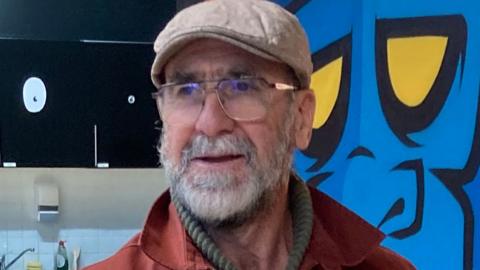 Eric Cantona, wearing glasses, a beige cap and a red jacket, stands in front of a painting of a blue monkey with yellow eyes