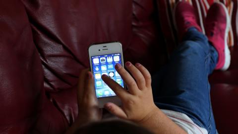 An over the shoulder shot of a child as they scroll through the home screen of an iPhone 4 while lying on a sofa.They are wearing a white top with thin red stripes, blue jeans, and pink trainers with white soles.