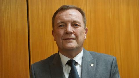 Council leader Tony Johnson, a man wearing a grey suit and white shirt with a dark-coloured tie. He has a pin badge on his lapel, and is stood against a wood-panelled background.