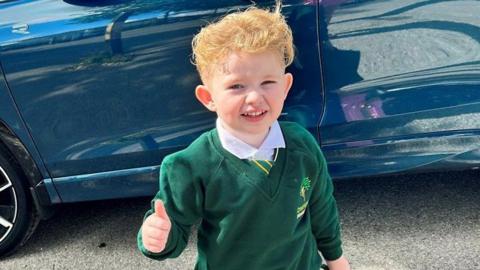 Peter Maughan, a four-year-old boy with short, blond hair, is standing by a blue car wearing a school uniform. He has a white shirt, a green and yellow tie and a green jumper. He is holding a green schoolbag and is making a thumbs-up sign.