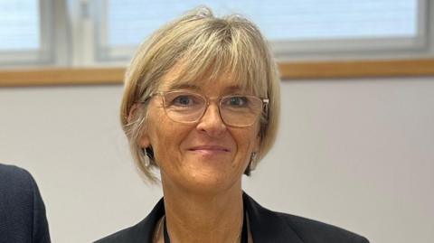 A woman with a blonde bob wearing a navy blazer and clear glasses smiling for the camera standing in an office room. The shoulder of another person is visible on the left.