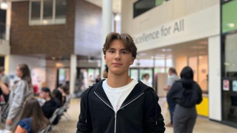 A boy standing in a school hall, he is wearing a black zip up hoodie top and a white t-shirt. 