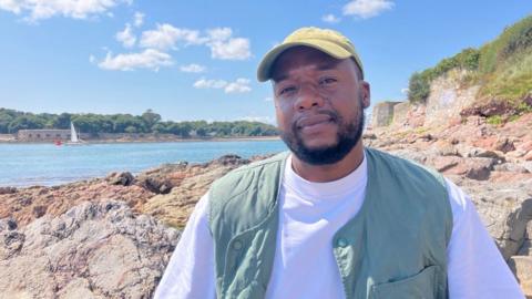 A picture of a man smiling at the camera. He is wearing a white shirt and a green gilet. He is wearing a green cap and is stood in front of rocks and the seafront. A boat can be seen in the distance.