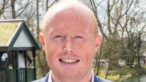 A man with ginger hair and wearing a suit, smiles while he stands outside. There are some trees behind him and a white and black hut to the left.