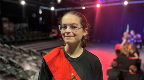 A child wearing a black shirt and red sash across their body. They are stood in front of a theatre auditorium.