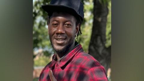 A picture of Kunta Ceesay wearing a red and black checked shirt and a black flat cap. He is smiling at the camera, standing in front of trees on a sunny day.