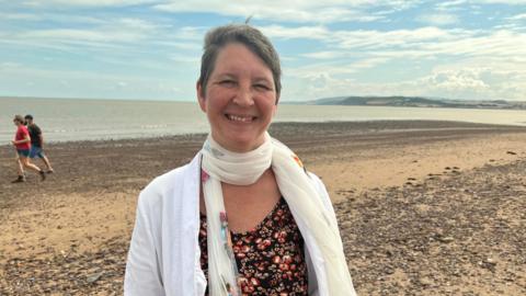 Reverend Chloe Kingdon wearing a white scarf and a red and black top. She has a white cardigan on too. She is standing on a beach. 