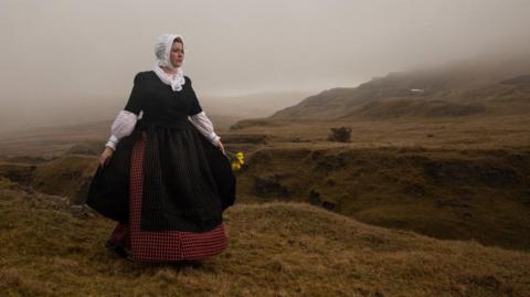 A woman is stood on the rolling hills in Wales. It is a grey, gloomy day with mist coming over. The hills are a mossy green. She is wearing traditional Welsh clothes, a black dress over a red skirt with a white undershirt, you can see the lower half of the sleeves. She has a white head wrap on as well and is holding daffodils. 
