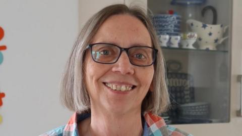 Hilary Stobart is smiling at the camera, standing in front of a display cabinet full of china. She has bob-length fair or greying hair and is wearing dark-rimmed spectacles and a colourful checked shirt