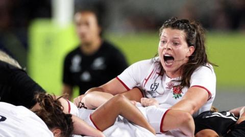 A female rugby player, with long dark hair and wearing red-edged white England rugby shirt, appears to be shouting at team-mates who are locked together and pushing.