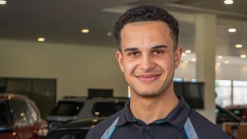 Tyrell Cambridge has short brown hair and is wearing a navy and silver polo shirt. He is standing in a car showroom which has several vehicles and large windows.