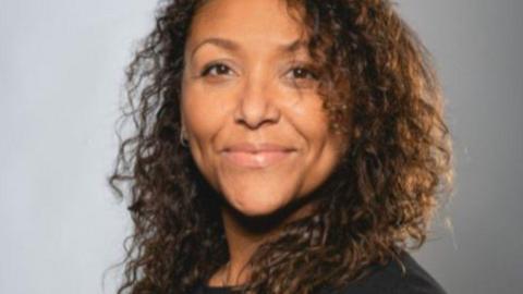 A headshot of Lynette Leith. She has long curly dark hair and is looking towards the camera, smiling.