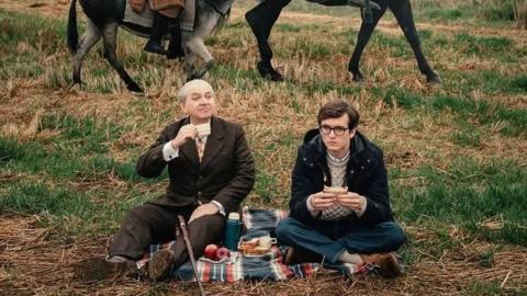 Luis Gnecco in brown suit and Fionn Whitehead in dark duffel coat are having a picnic, sitting on a rug in a field. The lower legs of two donkeys can be seen in the background.