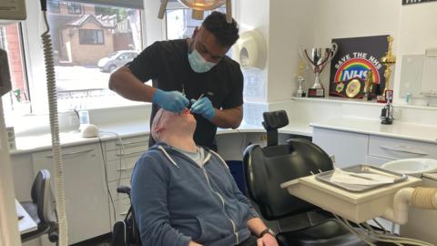 A man in a blue hoodie has his mouth open and is being examined by a dentist, wearing a mask and gloves and a black top.  On the wall of the examination room is a sign with 'save the NHS' and a number of trophies and boxing belt.
