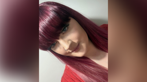 A woman with dark red straight hair takes a selfie while wearing a red top.