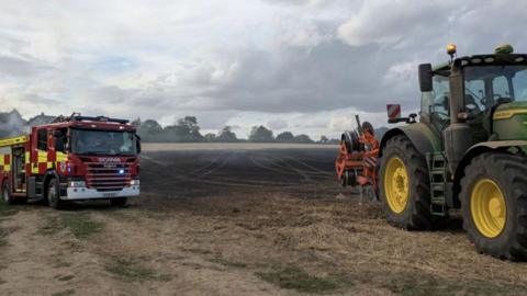 A stubble field which has a red and yellow fire engine on the left and a green tractor with large black wheels with yellow hubs on the right. In between is a blackened stretch of stubble, still slightly smoking, and beyond that are trees and a cloudy sky.