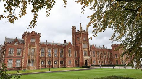 A photo of Queen's University main building (the Lanyon building), it's red bricked with smaller yellow masonry around the windows, which consist of 3 floors of windows and larger windows on the top floor. A central tower is the main entrance with four floors of windows. The building sits on a patch of green grass with a walkway to the main entrance. 