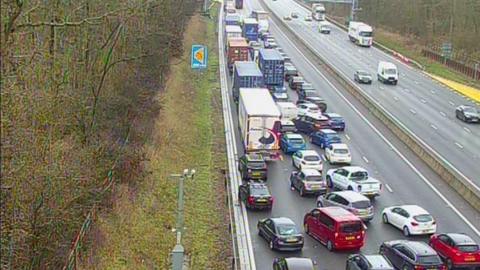 A CCTV image of a dual carriageway. On the left hand side are a large number of cars grouped together, with some of the vehicles moving into different lanes.  There are a number of lorries too. On the right hand side is four vehicles travelling on a much emptier road. Either side of the carriageway are trees and greenery. 