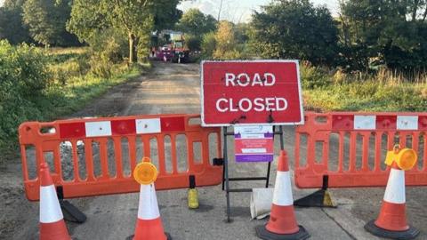 A red sign that reads road closed sits in front of orange barriers and behind orange cones. Machinery can be seen in the background working on a burst water main. 