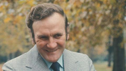 Don Revie with combed brown hair, smiling, as he walks in a park. He is wearing a grey blazer, blue shirt and navy tie. There are autumn trees behind him. 