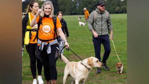 A group of people walk through a field on a charity walk. A woman with a greyish blonde bob holds her fist up and smiles as she walks a yellow Labrador, while a man beside her with a ginger beard and a cap on walks a sausage dog, who is wearing a jumper.