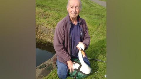 John Stewart kneeling on grass next to a canal with a swan between his legs. John has thinning grey hair and is clean shaven while he wears a mauve fleece over a dark blue jumper with a blue polo shirt collar visible underneath and has blue trousers. The swan has white feathers and an orange beak and  a green "vest" wrapped around it to protest people while transporting it. 