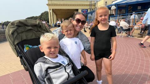 Tesni Parry pictured in sunglasses on Barry Island Promenade crouching by a buggy with a blond toddler boy inside and two other young blond children by her side