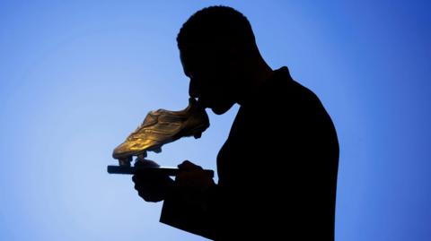 Silhouetted Mbappe holds and kisses trophy against a dramatic blue gradient background.