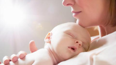 A baby sleeping on his or her mother's chest. The mum's chin, nose and lips can be seen with her chin gently resting on the baby's head. She wears a light top and has a blonde bob. 