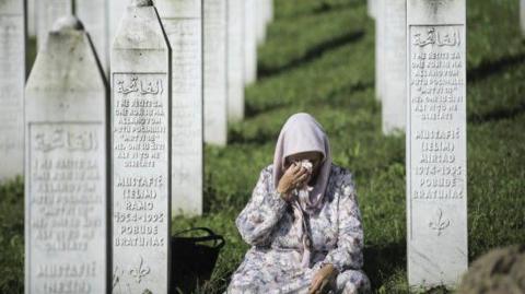 A woman is sat on the ground and weeps at a memorial to the victims of the Srebrenica massacre