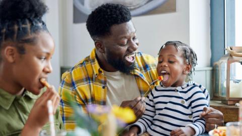 A father feeds a piece of fruit on a fork to his young daughter, who is sitting on his lap. Her older sibling is also sitting at the table