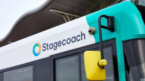A picture of the side of a bus - which says the word "Stagecoach" on it. It is a blue and white bus with a yellow wing mirror. There is a small camera next to the logo.