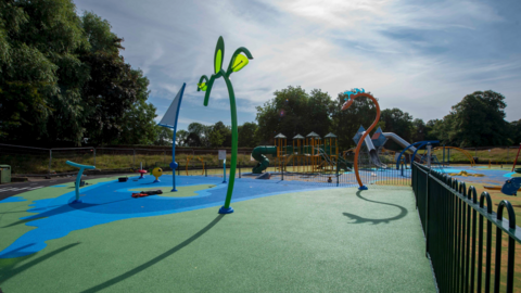 A water play area including structures that might be found on a children's playground. Trees are on the far left and railings are on the far right.