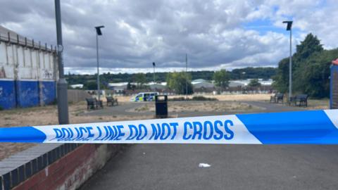 Blue-and-white police tape with the words "Police line do not cross". Beyond the tape, slightly out of focus, there is a litter bin and a circular path around a patch of grass. There are benches on either side of the path. On the left, there is a blue and white building with peeling paint. There are streetlights on poles. In the distance there is a police van.