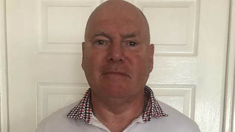 Philip Miller, who has a bald head and is wearing a white shirt with a dark red and black checked collar, is standing in front of a white door in a house. He looks impassive. 