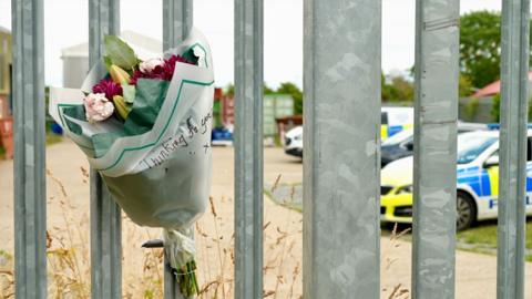 A close up of a bunch of flowers that has been strapped to the metal gate of the factory. It says Thinking of You on the bouquet and the name has been blurred out. Through the silver bars you can see a police car and police van parked alongside a black car. 