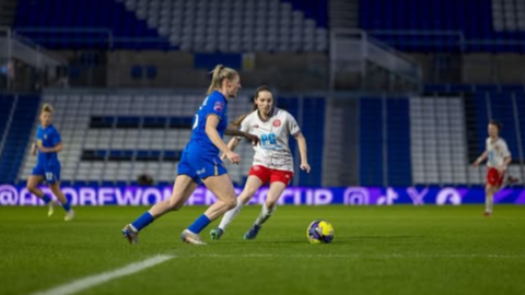 Players from Birmingham City and Chatham Town are on the pitch, with other team members in the background. There are football stands behind them.