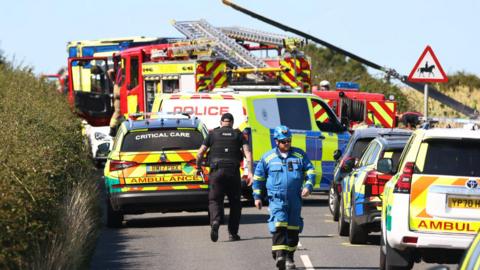 Two emergency service workers walk between emergency vehicles, surrounded by fields.