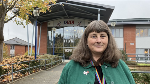 A woman with thick shoulder length brown and grey hair with a fringe. she is wearing a green jacket with an RNC and Poppy pin and she is wearing a rainbow lanyard around her neck. she is standing in front of a glass building with the letters RNC in blue and red.