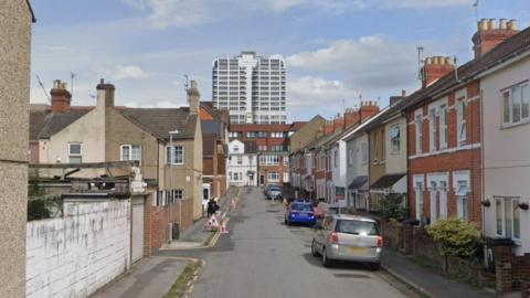 A view of a part of Tennyson Street in Swindon with a highrise building in the background, a few parked cars on the right and two cones and a roadworks sign on the left