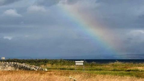 Plenty of cloud and a rainbow over East Lothian, Scotland. There is yellow grass in the foreground and tall cream-coloured buildings in the distance.