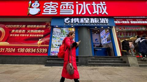 A woman wearing a red coat walks past a Mixue Ice Cream & Tea store while using her smartphone on 22 February, 2025 in Chongqing, China.