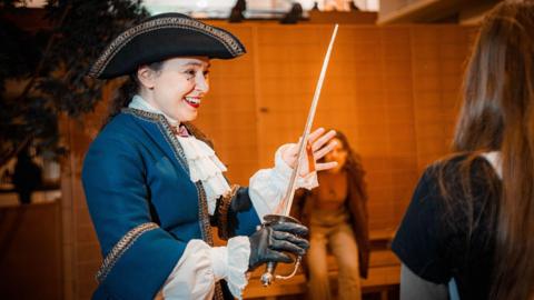 A woman in a historical French costume of a tricorn hat, blue jacket and white shirt smiles as she holds a sword.
