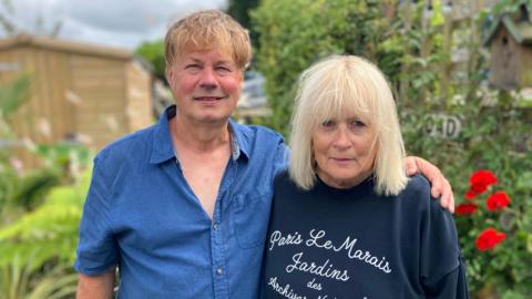 Marc Slegg is wearing a blue button up top. Lesley Raache, with blonde hair, is wearing a blue jumper. Marc has his arm around Lesley. Behind them is the blurred background of the garden with a wooden shed and flowers.