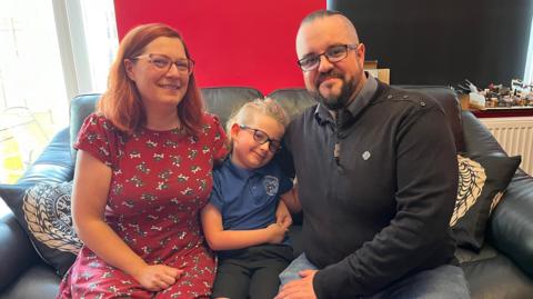 Seven-year-old Hadrian sits in the middle of its parents on a dark coloured sofa. The child is wearing their school uniform which is made up of a blue polo shirt and black shorts. To the left his mother Wendy is wearing a red dress with a grey and white pattern. She has shoulder-length ginger hair. To the boy's right is his father Robert who is wearing a dark navy jumper and blue jeans. He has a long braided beard. All three are wearing glasses.