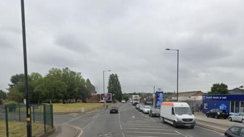 Golden Hillock Road in Birmingham. A general picture on a grey day with a white and orange van and a row of cars on the lane coming towards the camera and a single black car on the other side. The left-hand side of the picture shows an area of green grass and trees and some green fencing. On the left side is a garage. 