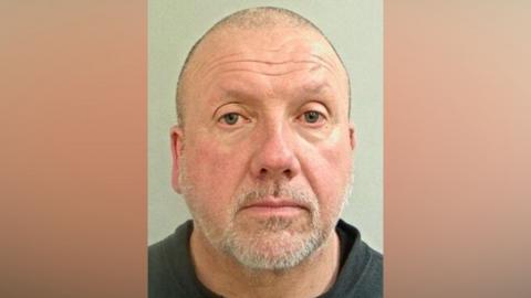 A mugshot of a middle aged man. He has a shaved head and a short grey beard. He is wearing a black T-shirt and black sweater jumper. 