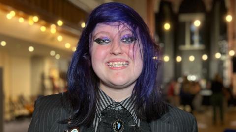 Lexie has dark, shoulder‑length hair and stands in a large hall decorated with string lights. They are wearing a dark pinstripe jacket with multiple badges and a ruffled shirt. Rows of chairs and a tall arched structure are visible behind them.