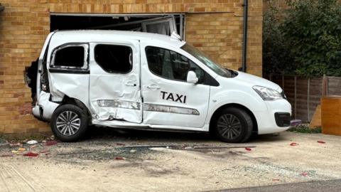 A dented white taxi embedded in the side of a brick house.